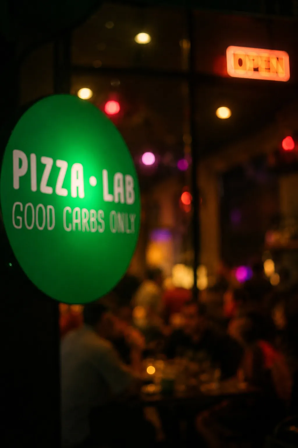 Green neon Pizza Lab sign and a lit OPEN sign at night, with guests inside